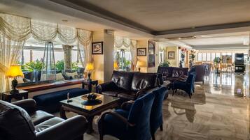 A spacious lobby with a leather sofa, blue armchairs, and a glass coffee table. There are two lamps on side tables, and a large mirror reflects the room.