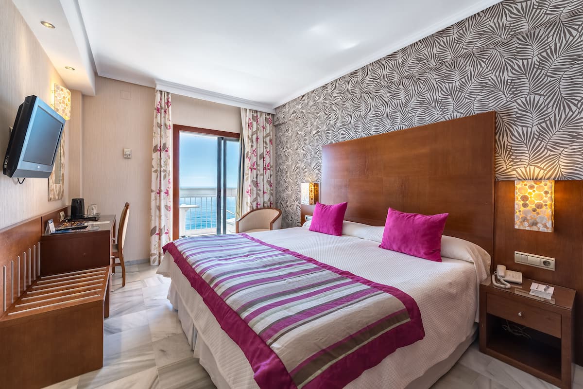 Een hotelkamer met een groot bed, een bureau, een stoel, een televisie en uitzicht op zee.