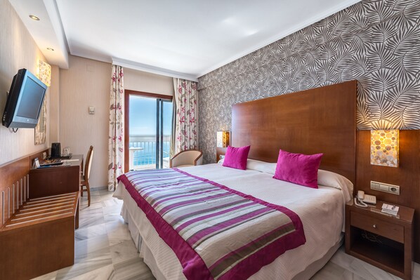 Habitación de hotel con una cama grande, un escritorio, una silla, un televisor y vista al mar.
