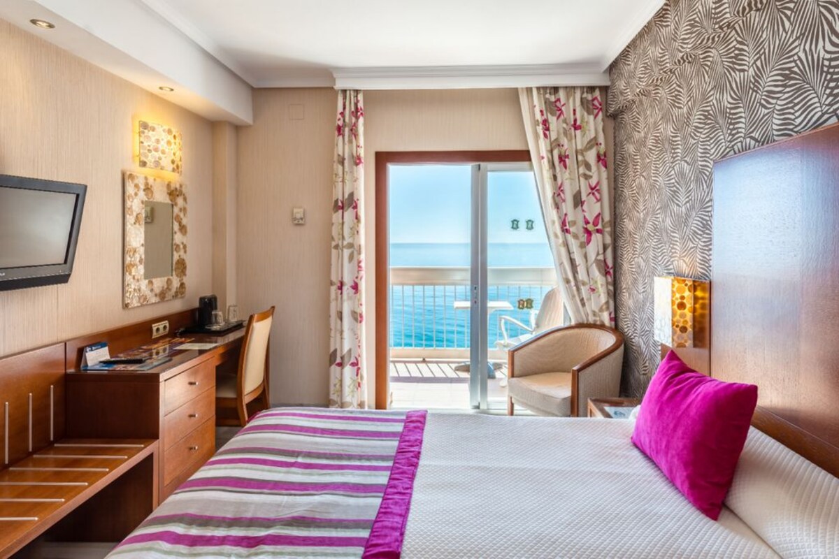 Habitación de hotel con cama, escritorio, silla, televisor y balcón con vista al mar.