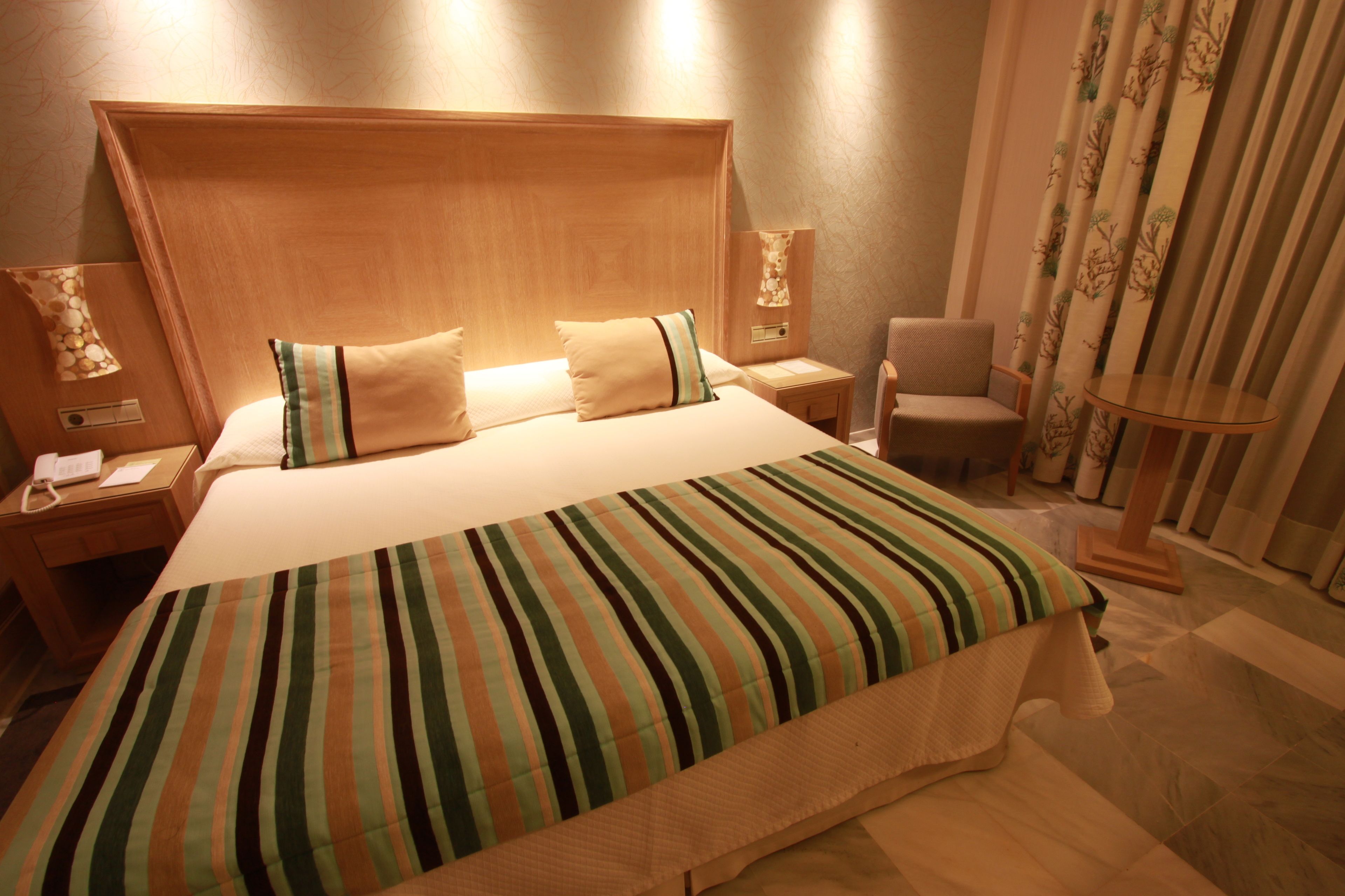 Habitación de hotel con una cama grande, cabecera de madera, dos mesitas de noche con lámparas, una silla y una cortina con estampado de árboles.