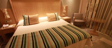 Habitación de hotel con una cama grande, cabecera de madera, dos mesitas de noche con lámparas, una silla y una cortina con estampado de árboles.