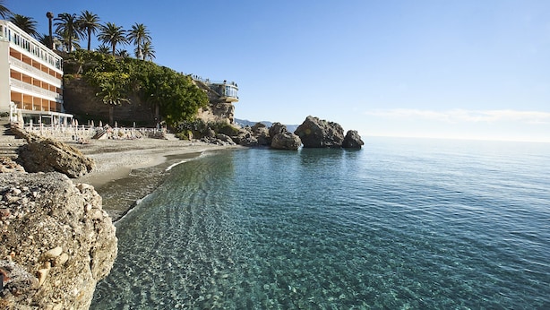 Una playa con agua cristalina, costa rocosa, palmeras y un edificio con balcones con vista al mar.