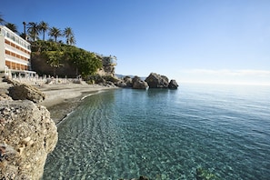 Una playa con agua cristalina, costa rocosa, palmeras y un edificio con balcones con vista al mar.