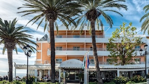 Un hotel con entrada central, mesas al aire libre, palmeras y cielo despejado.