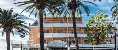 A hotel with a central entrance, outdoor seating, palm trees, and a clear sky.