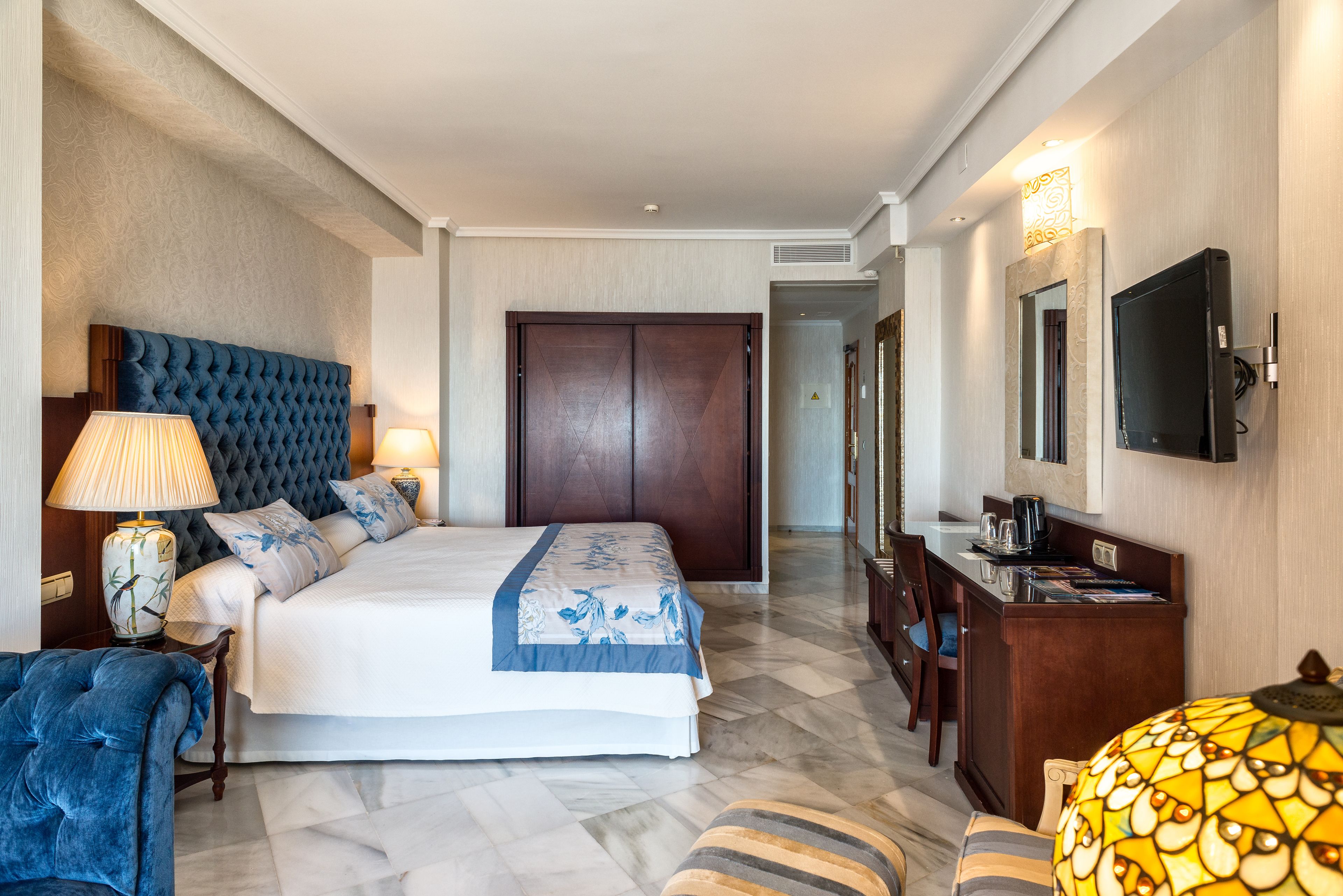 A hotel room with a large bed, a wooden headboard, a desk with a lamp, a television, and a patterned ottoman.