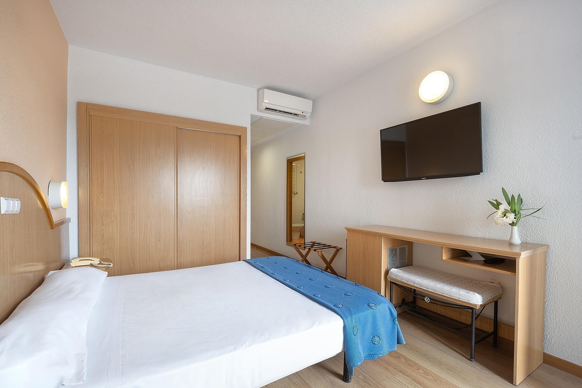 A hotel room with a bed, a desk with a chair, a television, and an air conditioning unit.