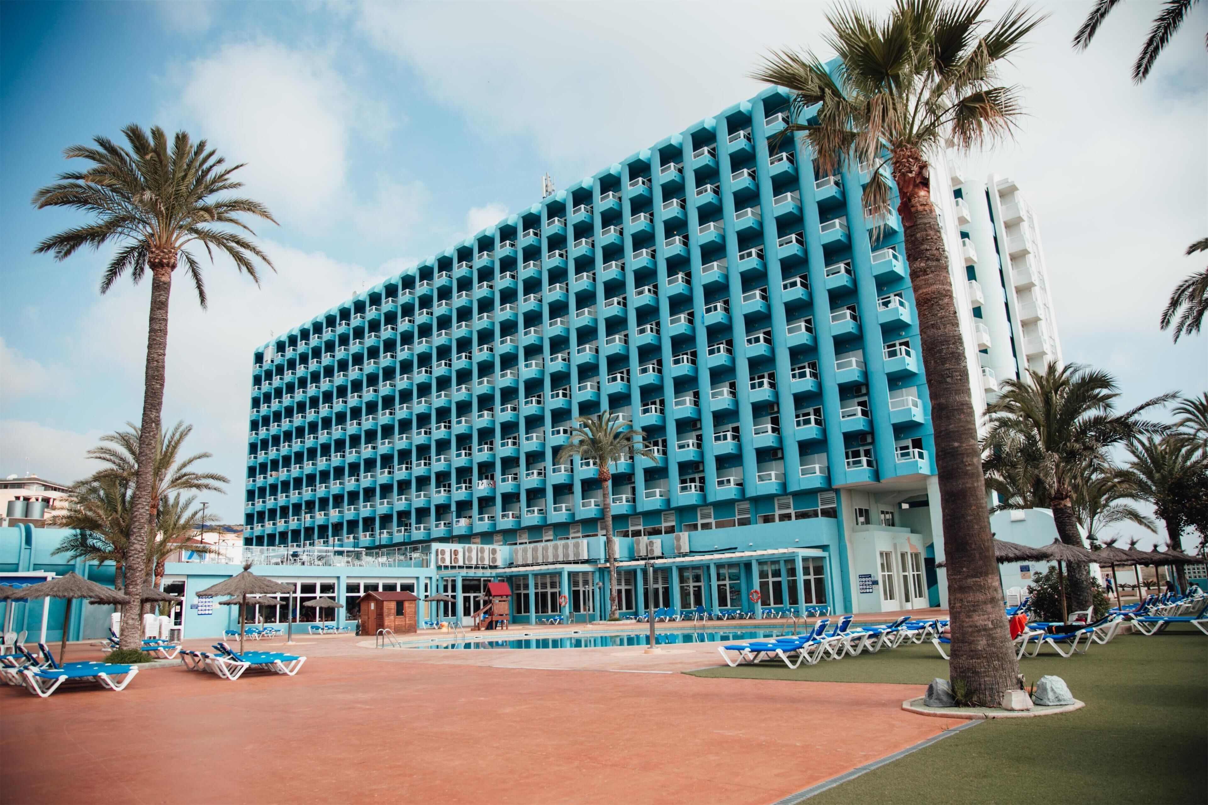 A large hotel with multiple floors, a pool area with sun loungers, and palm trees.