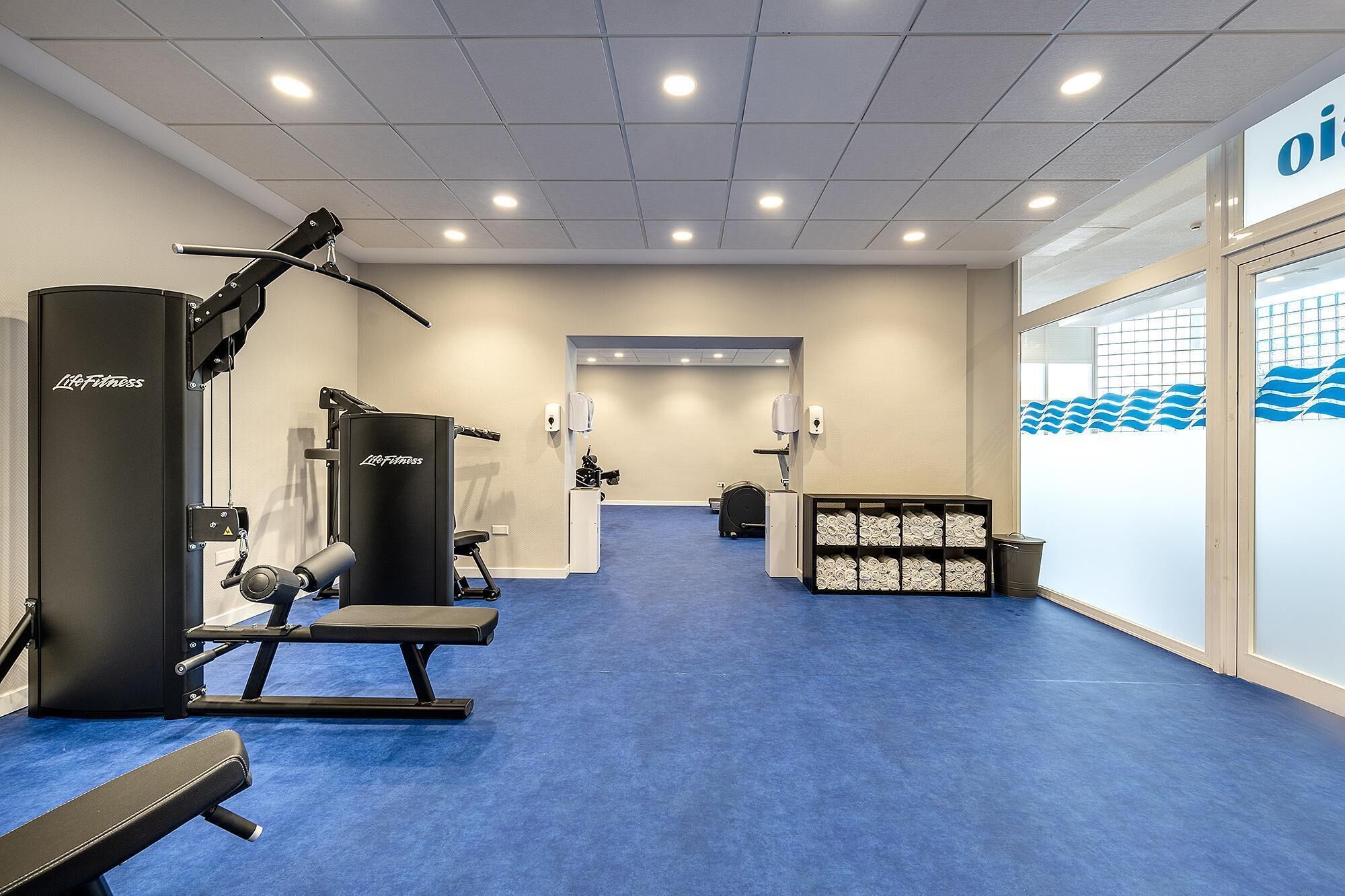 Et fitnessrum med træningsudstyr, et blåt tæppe og en væg med 'Life Fitness' logo.