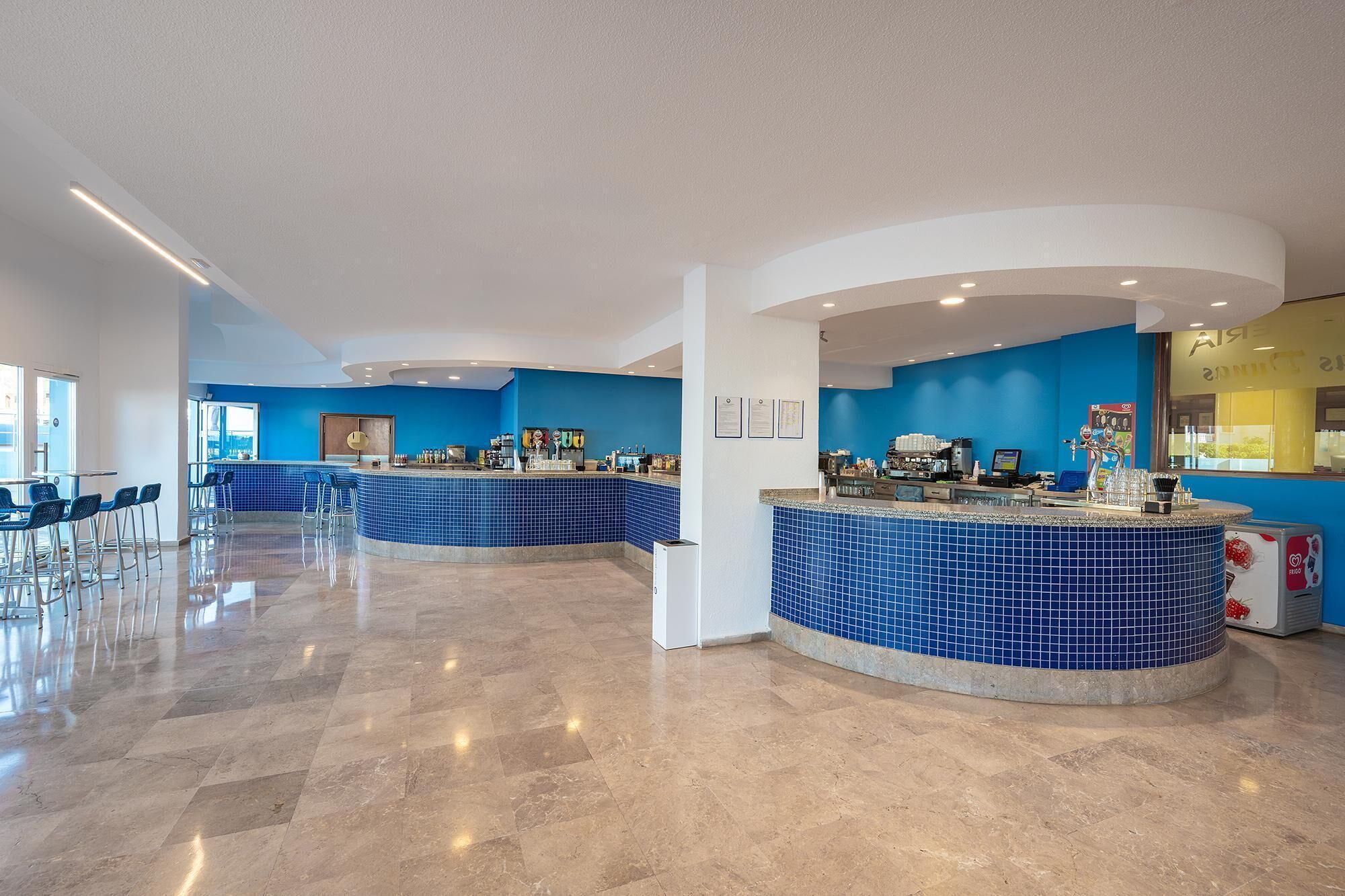 A modern hotel lobby with a blue-tiled reception area, a bar, and a seating area with chairs.