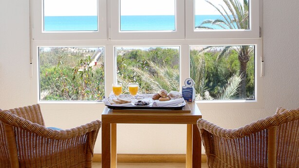 A dining area with a table and wicker chairs, overlooking a beach and palm trees through large windows. - Hotel Playas de Guardamar (Guardamar del Segura)