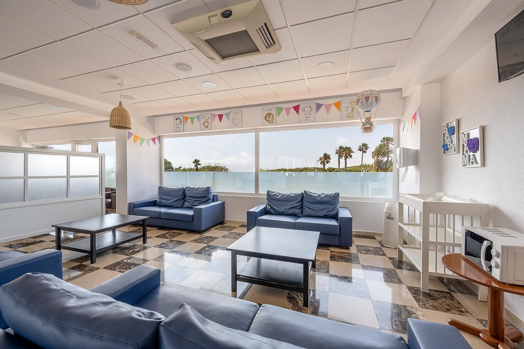 A modern room with two sofas, a coffee table, and a crib. Large windows offer a view of palm trees and a distant cityscape.