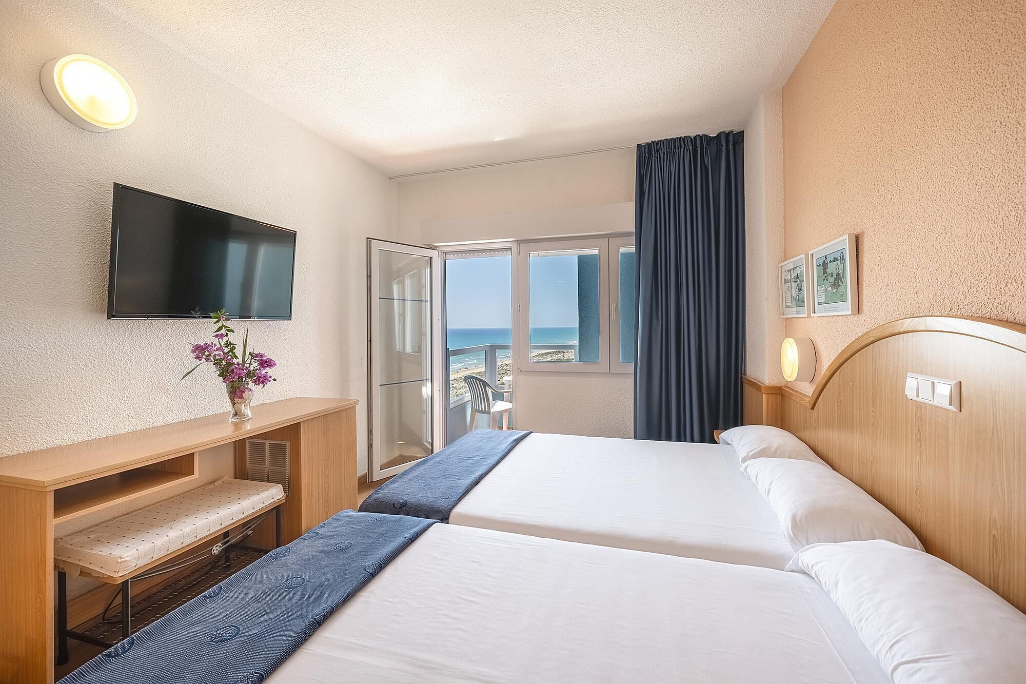 A hotel room with two beds, a TV, a desk, a chair, and a view of the beach.