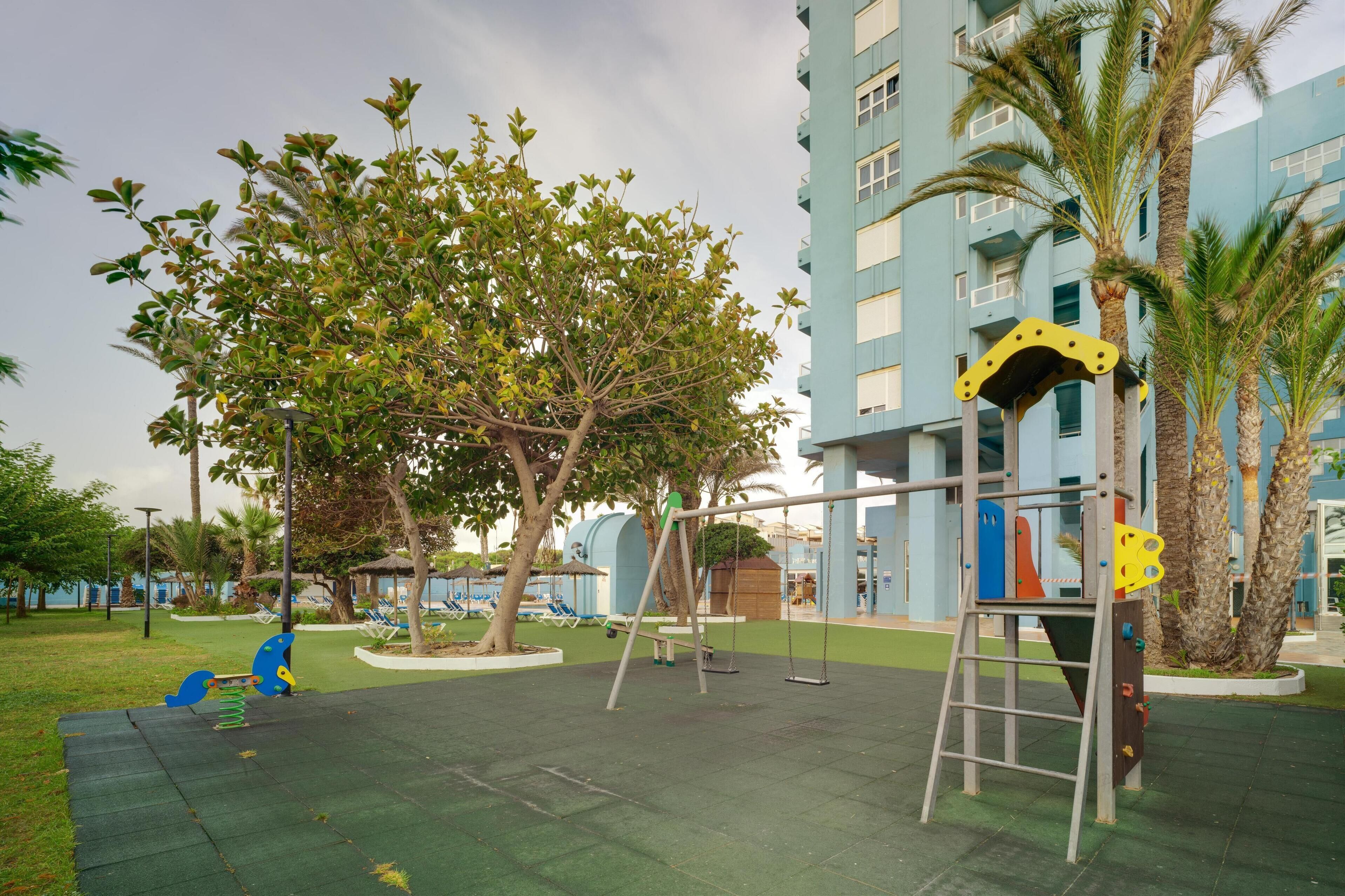 A playground with swings and a climbing structure, surrounded by palm trees and a building with balconies.