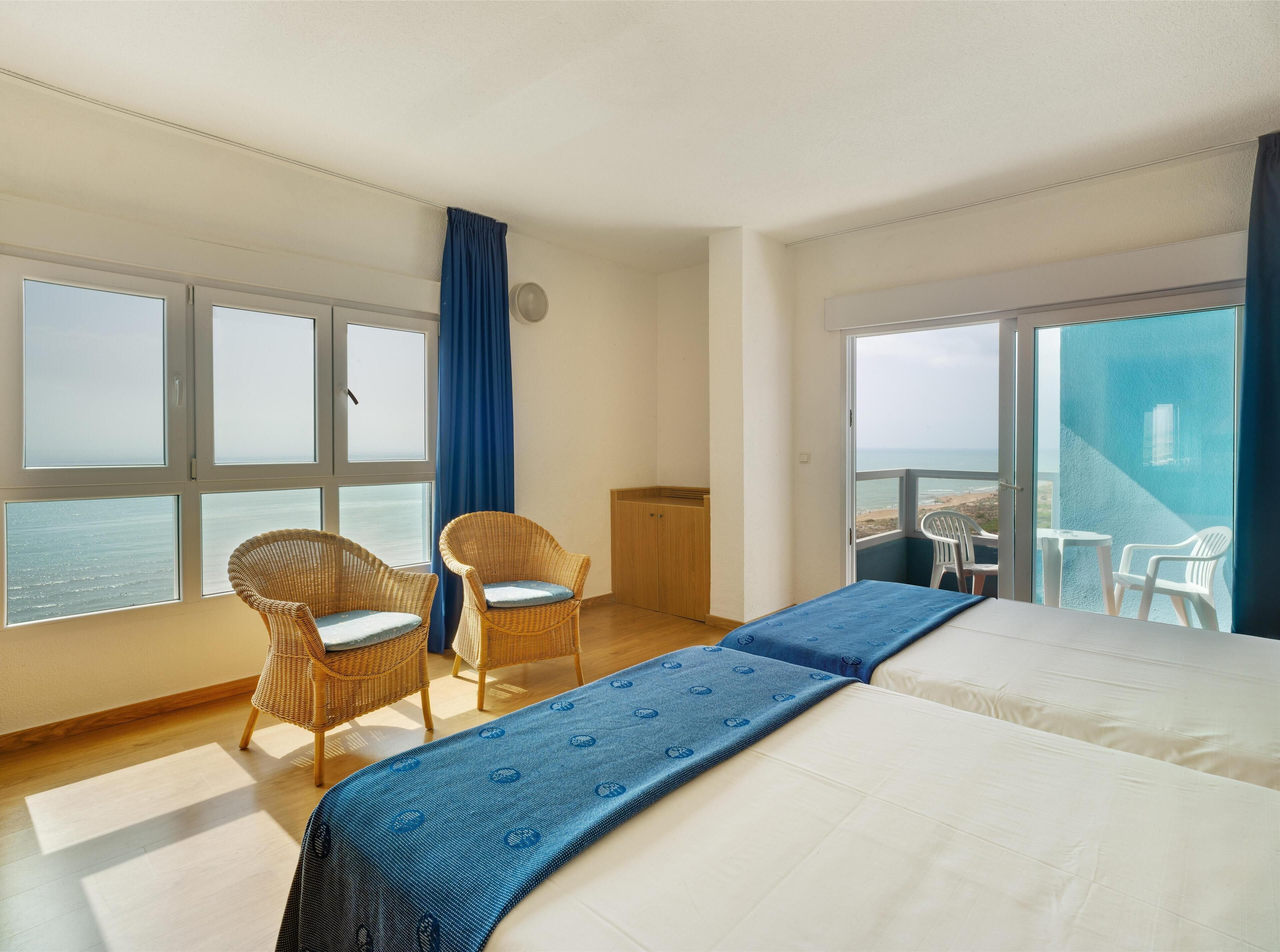 Habitación de hotel con una cama grande, dos sillones de mimbre, una mesita y un balcón con vistas al mar.