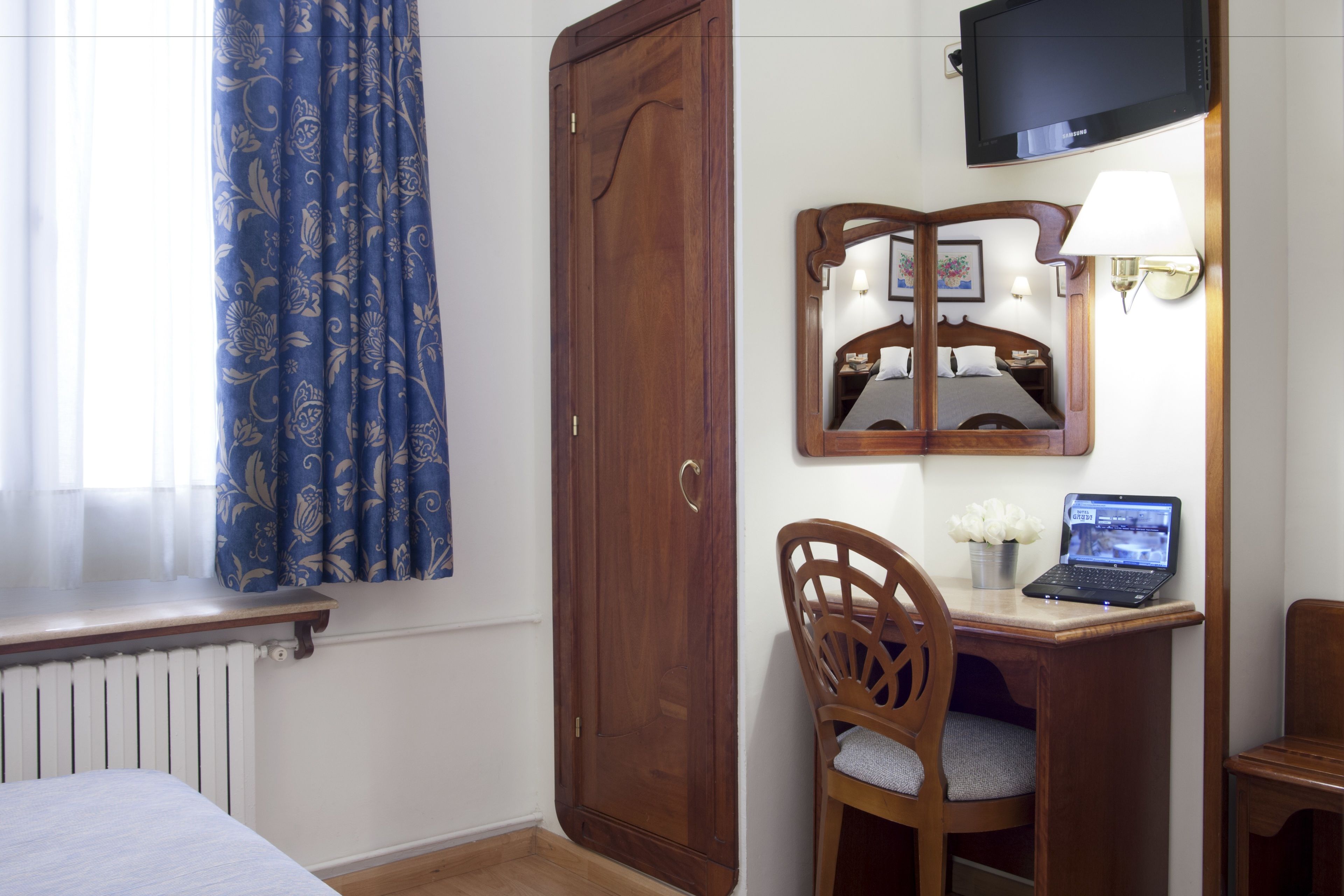 A hotel room with a wooden wardrobe, a desk with a laptop, a chair, a bed, a television mounted on the wall, and a window with curtains.