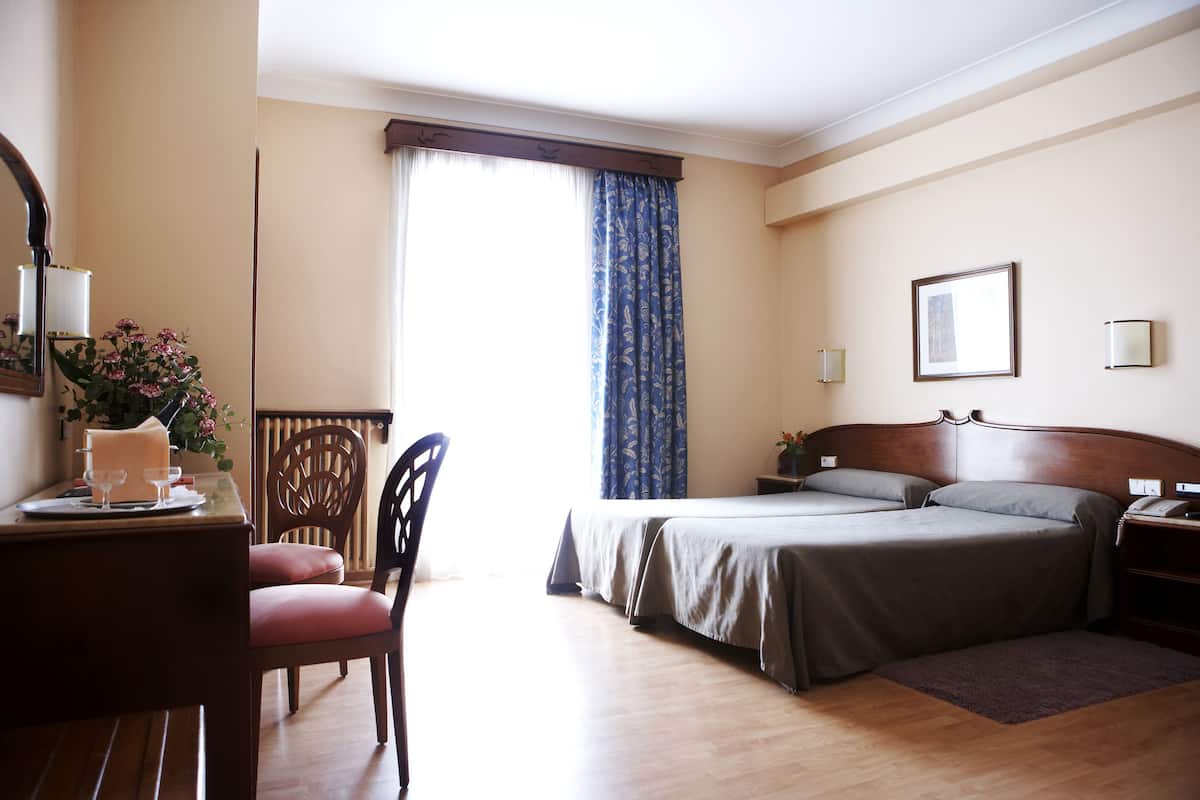 A hotel room with a bed, a desk with a chair, a window with curtains, and a bedside table with a lamp.