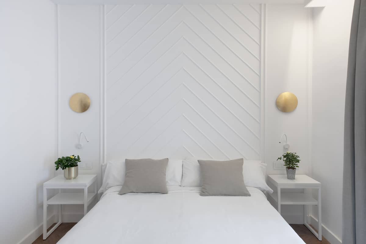 A modern bedroom with a white bed, two bedside tables with potted plants, and a geometric wall design.
