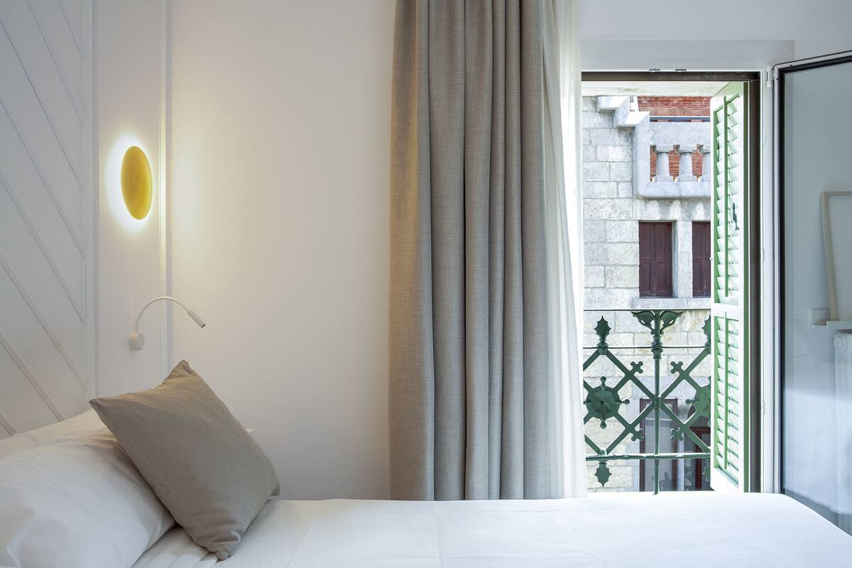 A hotel room with a bed, a nightstand, a wall-mounted light, and a view of the outside through a window with curtains.
