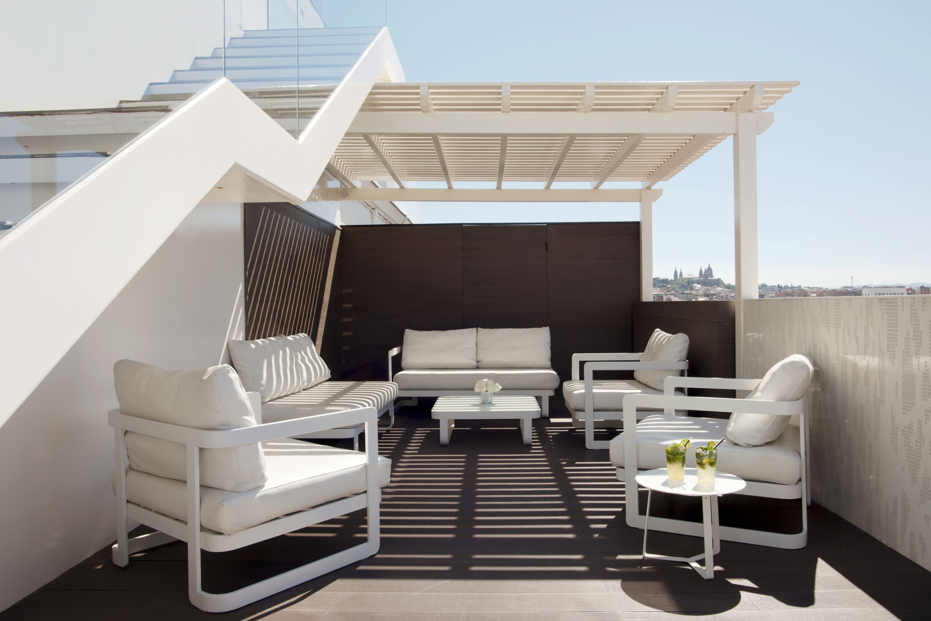 A rooftop terrace with white furniture, a small table with drinks, and a view of the city.
