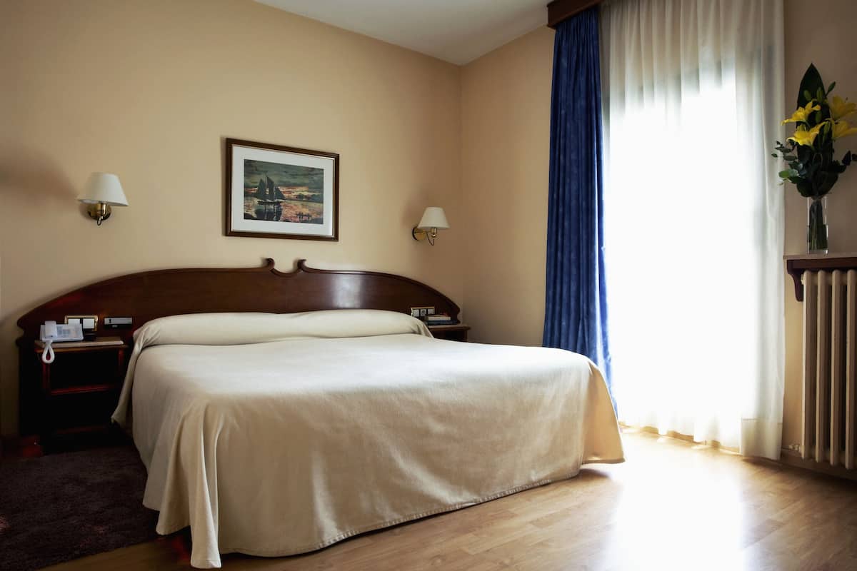 A hotel room with a large bed, two bedside tables, a nightstand, a framed picture on the wall, a vase with flowers, and a window with curtains.