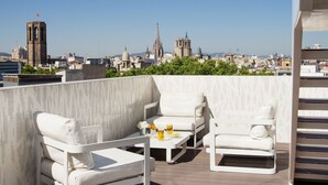 A rooftop terrace with white outdoor furniture, a table with drinks, and a cityscape view in the background. - Hotel Gaudí (Barcelona)