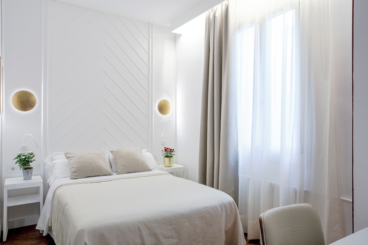 A modern hotel room with a large bed, a bedside table with a lamp, and a view of the outdoors through sheer curtains.