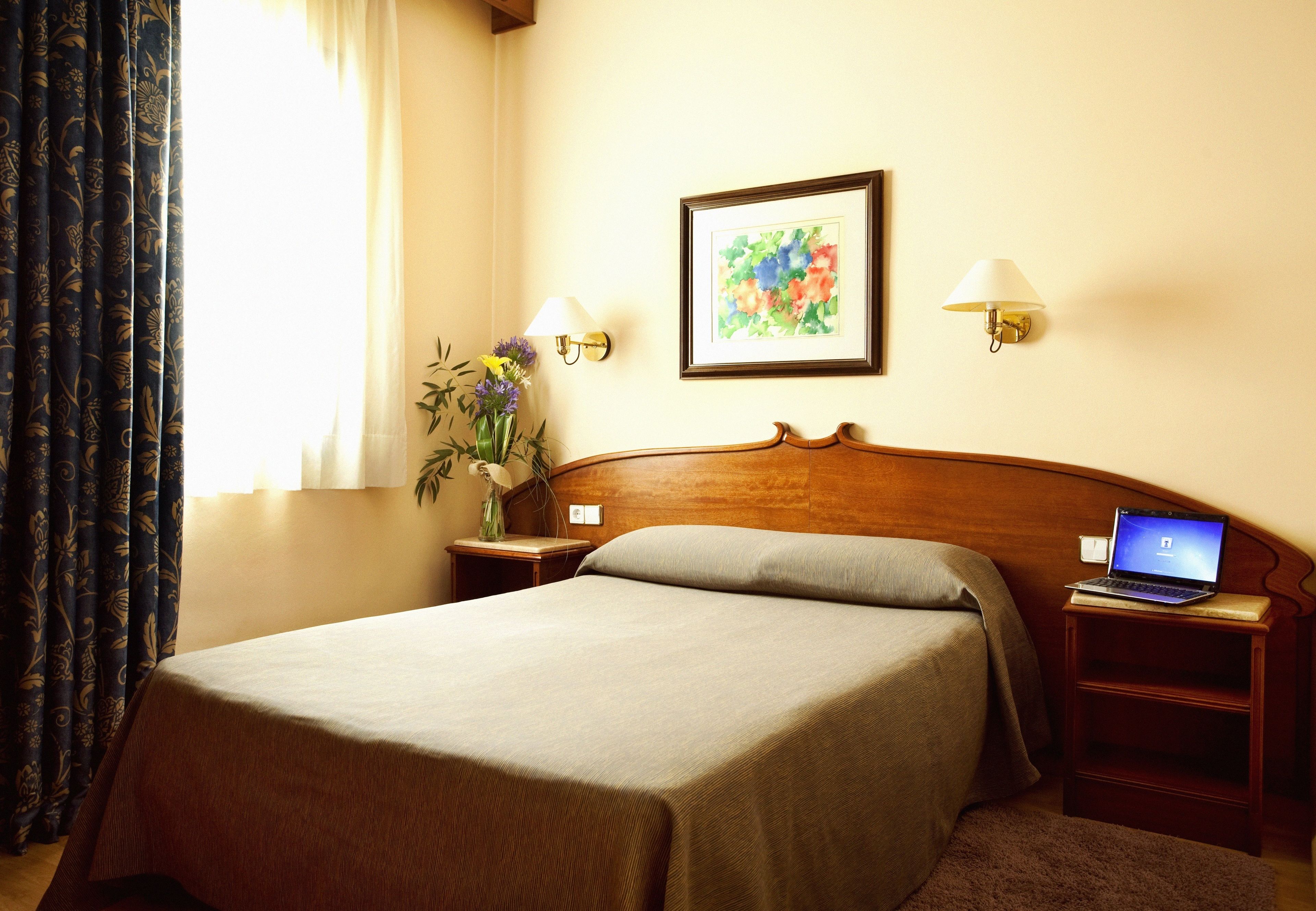A neatly made bed with a wooden headboard, a laptop on a bedside table, and a framed floral artwork on the wall.