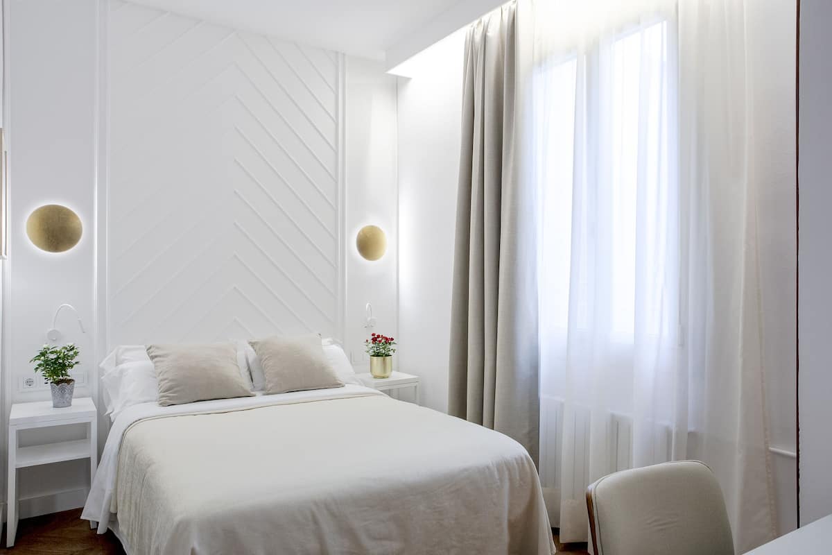 A modern hotel room with a large bed, a bedside table with a lamp, and a view of the outdoors through sheer curtains.