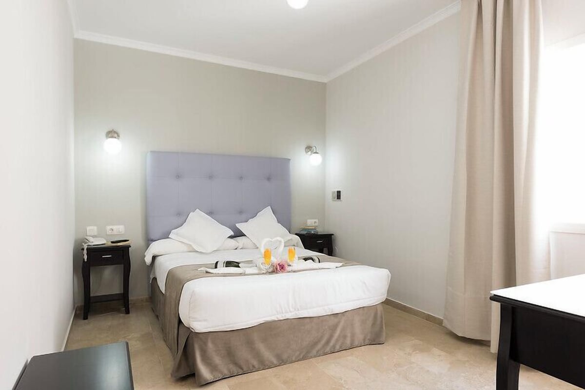 Una habitación de hotel con cama, mesitas de noche, escritorio y ventana con cortinas.