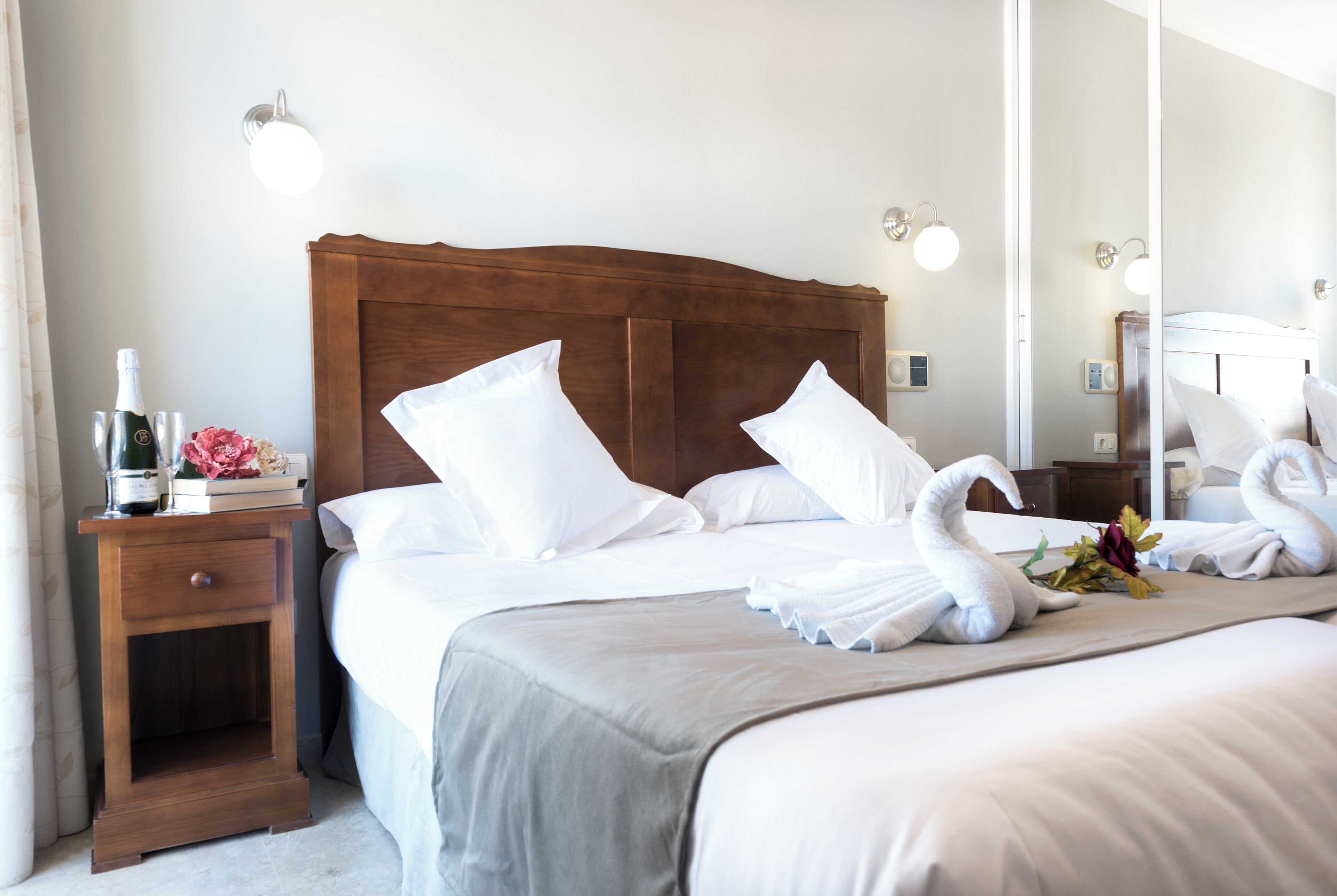 A neatly made bed with white linens and decorative swan-shaped towels, a wooden nightstand with a bottle and glasses, and a mirror reflecting the room.