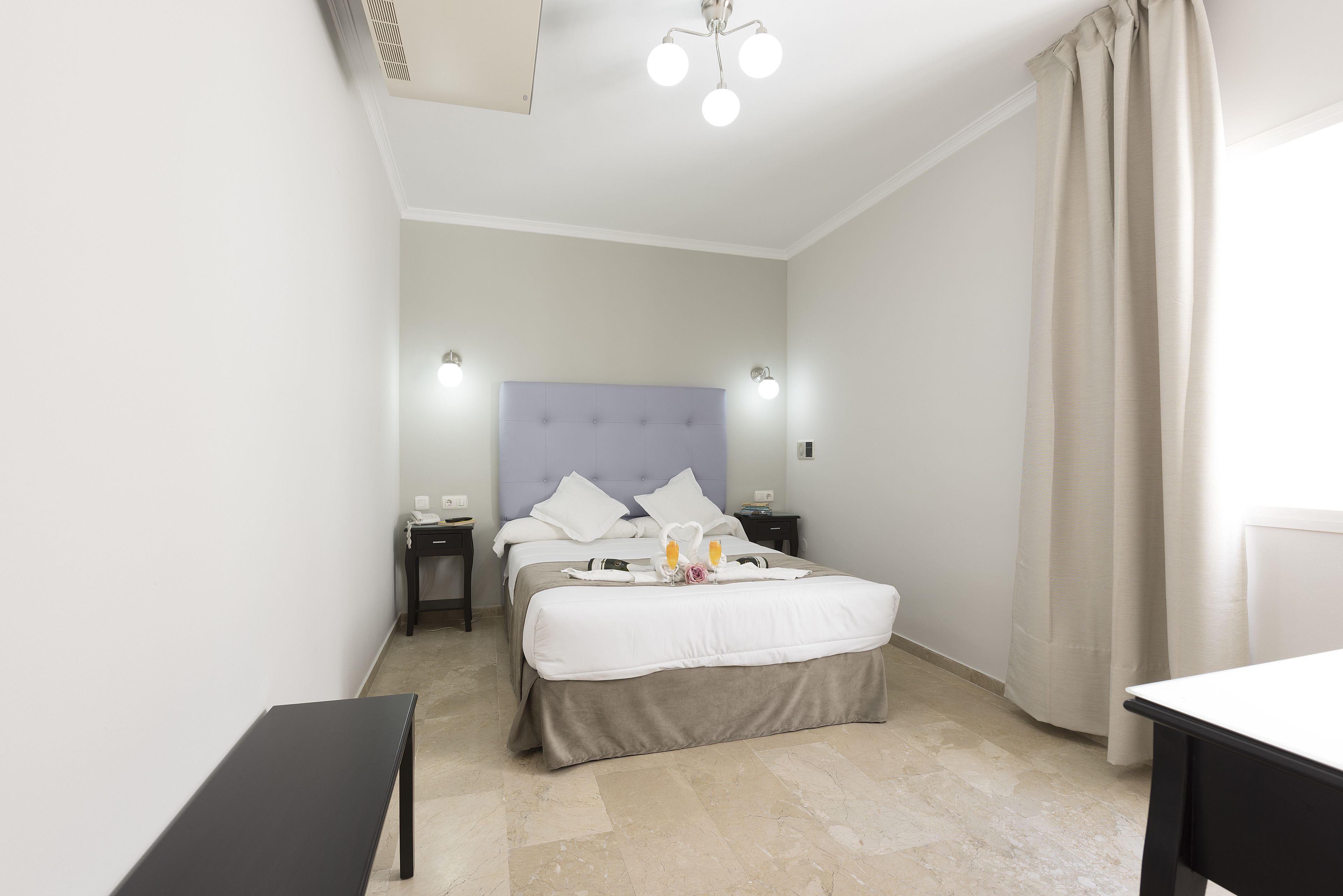 A hotel room with a bed, bedside tables, a bench, and a ceiling light fixture.