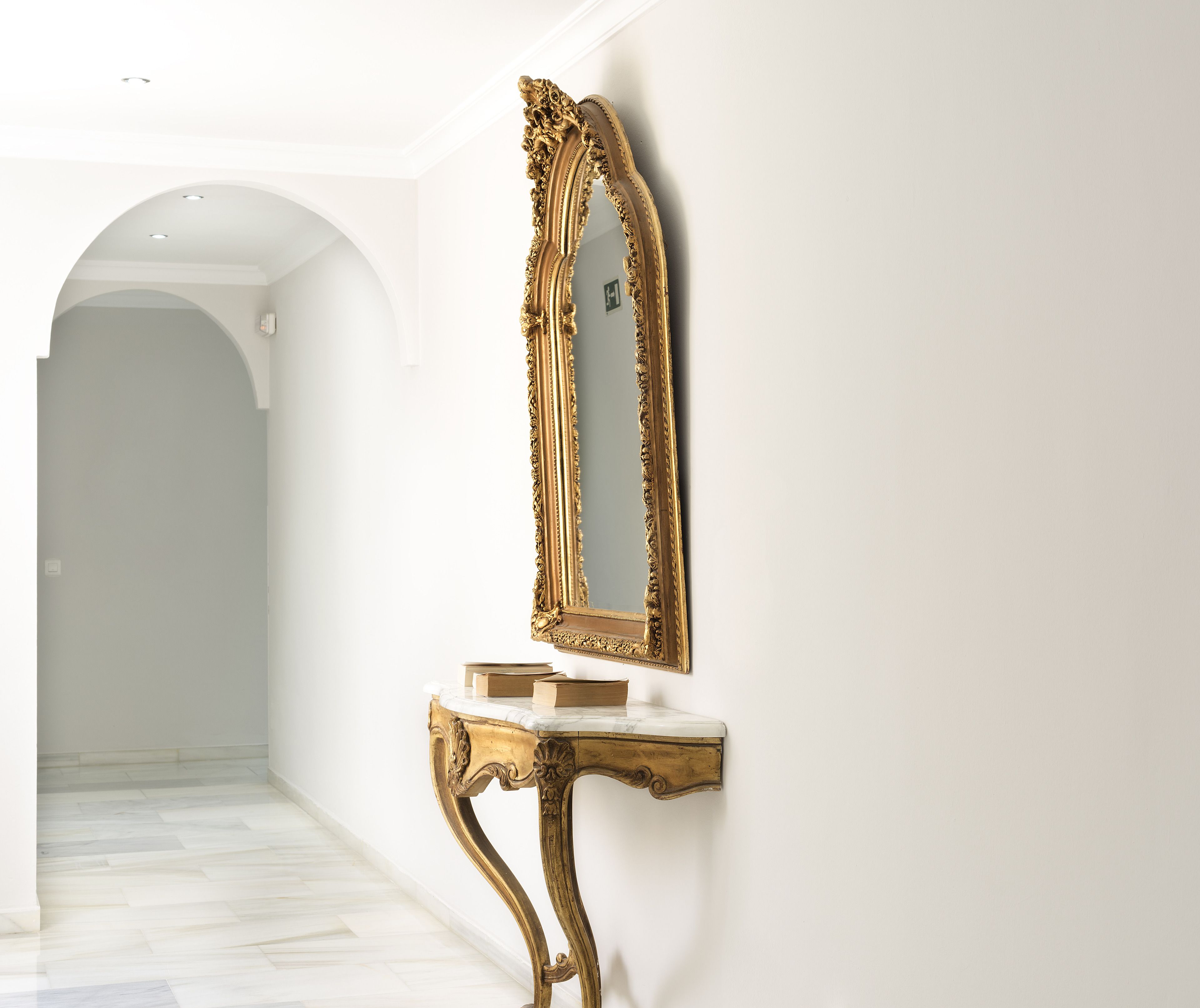 A hallway with a marble floor, a white wall, and a gold-framed mirror on a marble console.