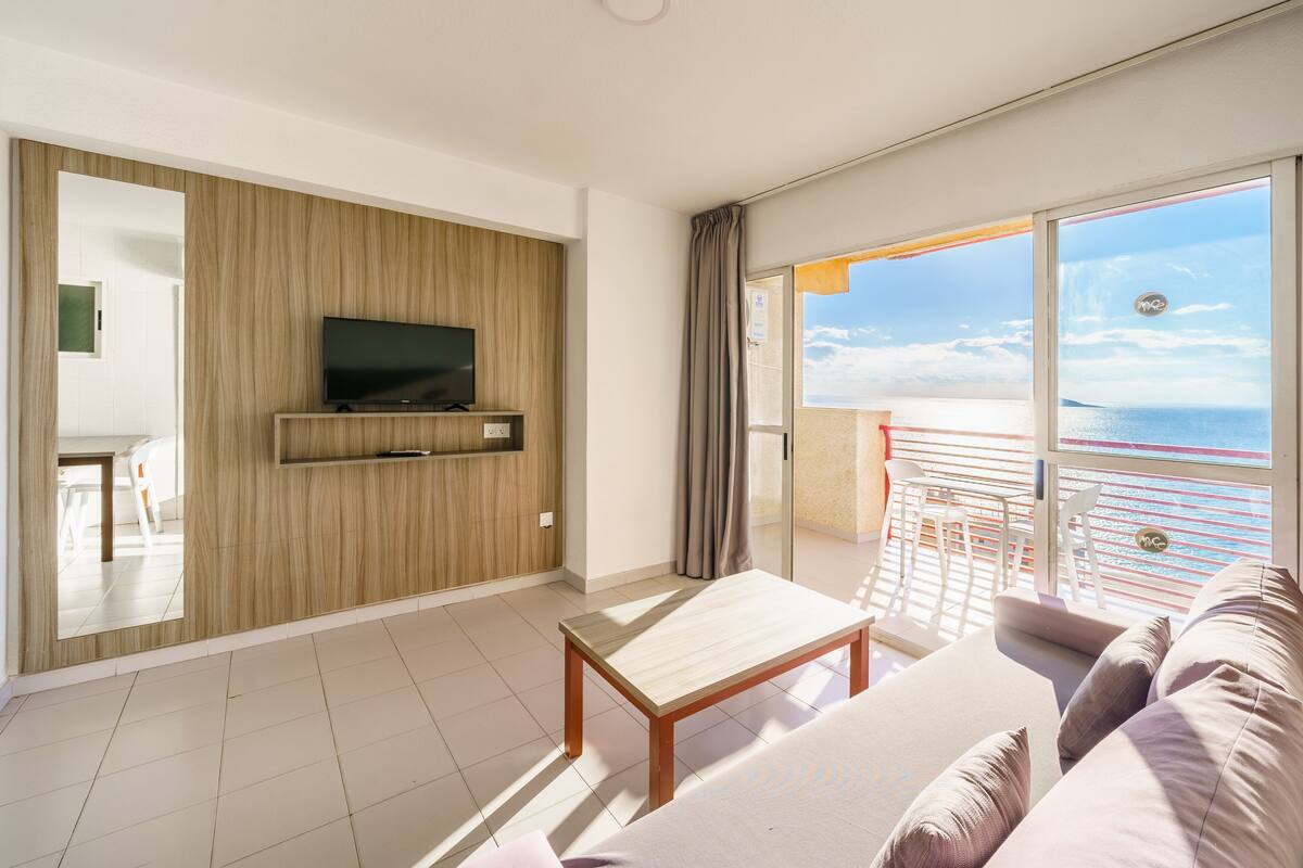 A modern hotel room with a TV mounted on a wooden panel wall, a balcony with a view of the sea, and a large sofa.