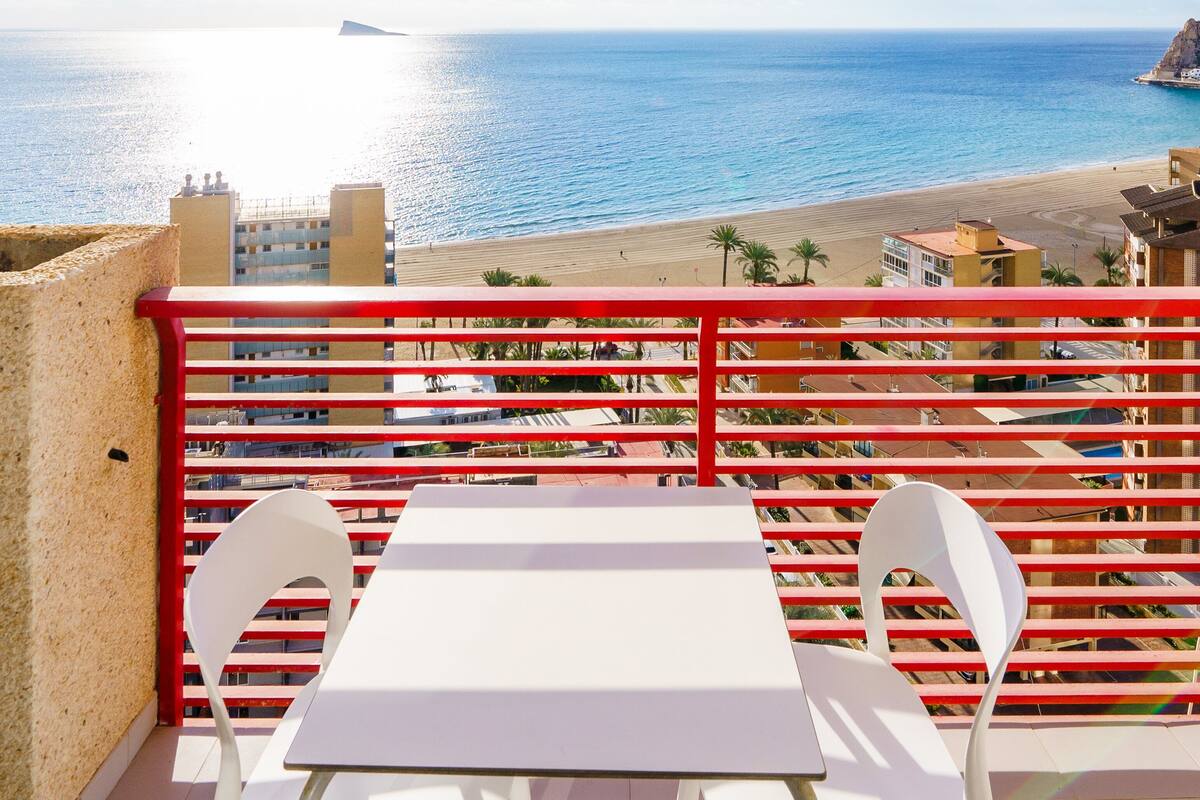 A balcony with a white table and two chairs, overlooking a beach with a clear sky and ocean.