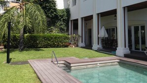 A swimming pool with a wooden deck, a covered patio area with outdoor furniture, and a white building with columns. - La Luna Blanca (Torremolinos)
