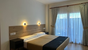 A hotel room with a wooden headboard, two bedside lamps, a bed with a beige and black bedspread, a nightstand, and a window with curtains. - La Luna Blanca (Torremolinos)