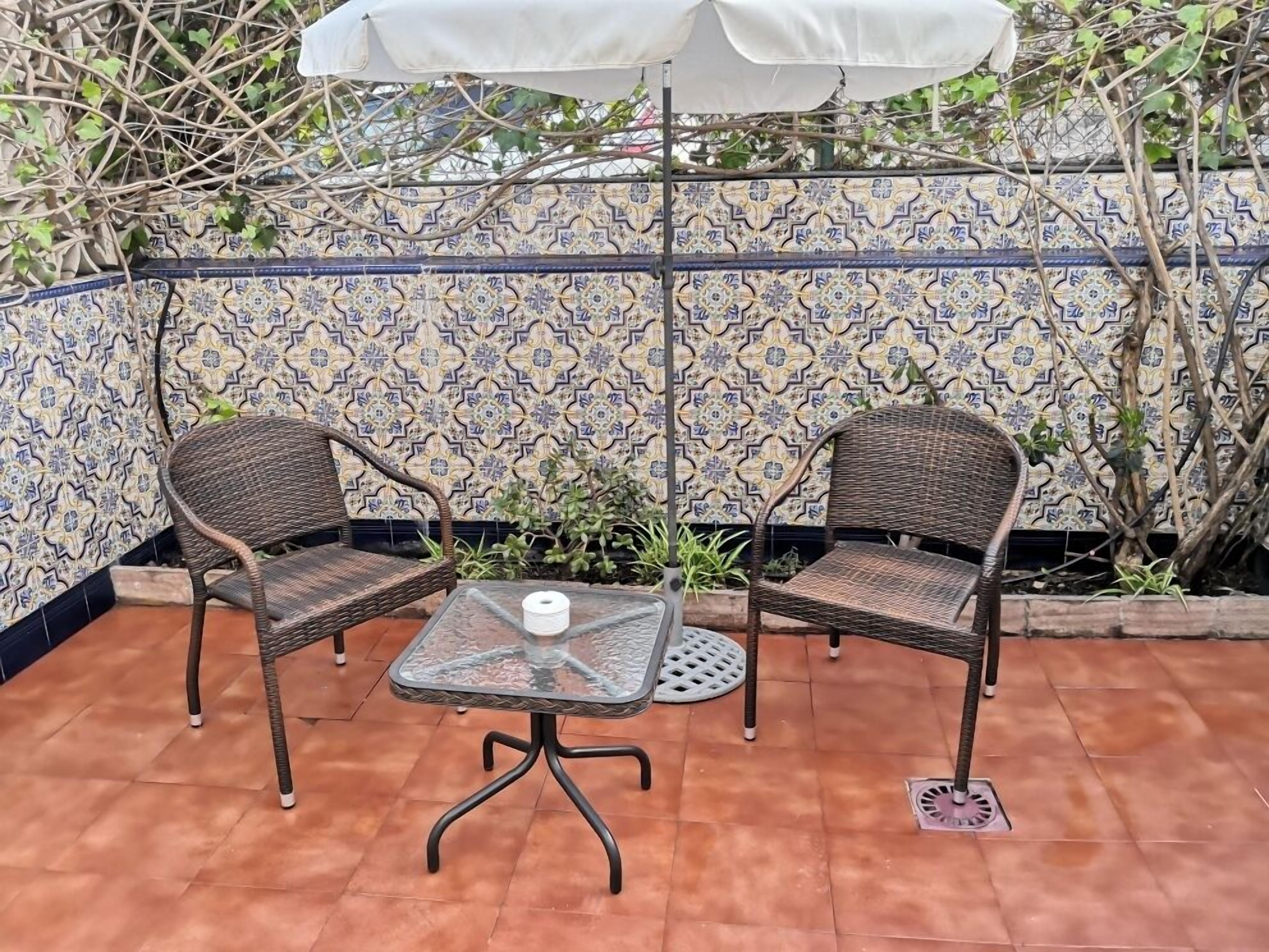 A patio with two wicker chairs and a glass table, an umbrella, and a tiled wall with a geometric pattern.