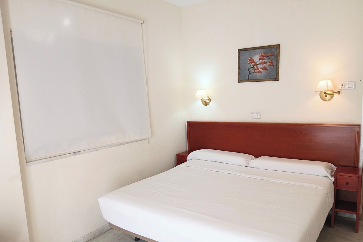 A hotel room with a single bed, a nightstand, a wall-mounted lamp, and a framed picture on the wall.