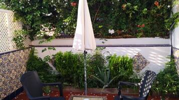 Un patio al aire libre con dos sillas negras, una mesa y un paraguas color beige. El espacio está rodeado por un muro blanco con azulejos azules y dorados, y hay varias plantas y flores al fondo.