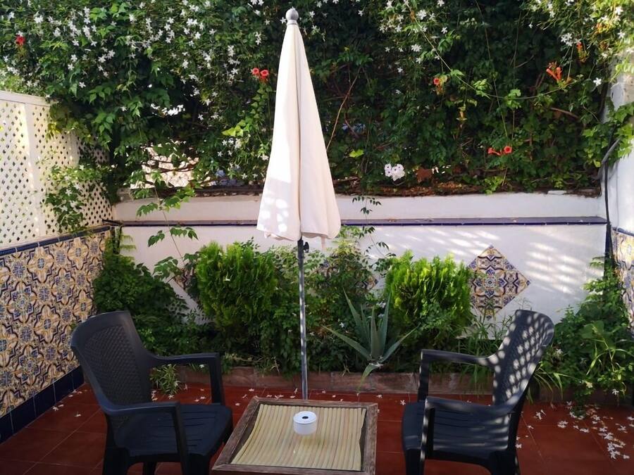 Un patio al aire libre con dos sillas negras, una mesa y un paraguas color beige. El espacio está rodeado por un muro blanco con azulejos azules y dorados, y hay varias plantas y flores al fondo.