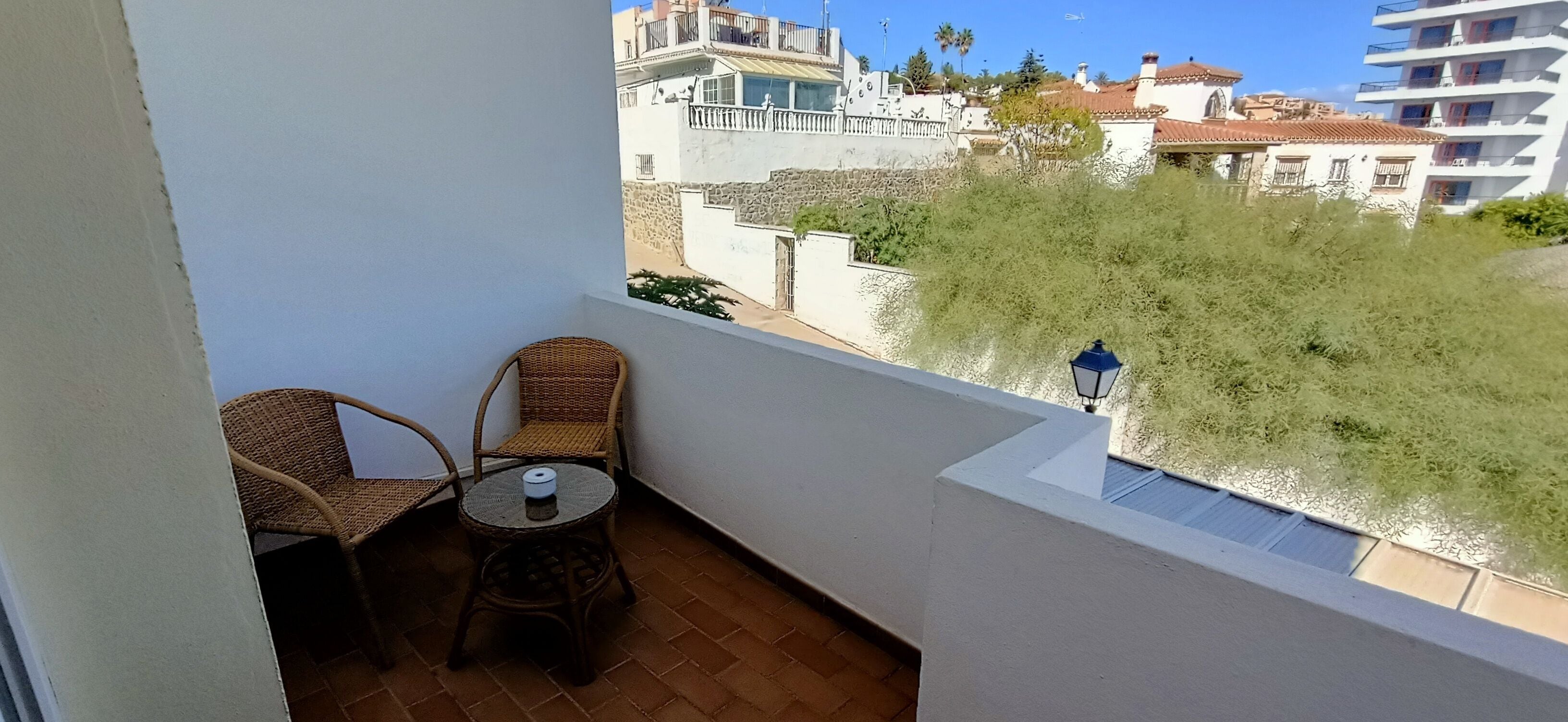 A balcony with wicker chairs and a small table, overlooking a residential area with buildings and greenery.
