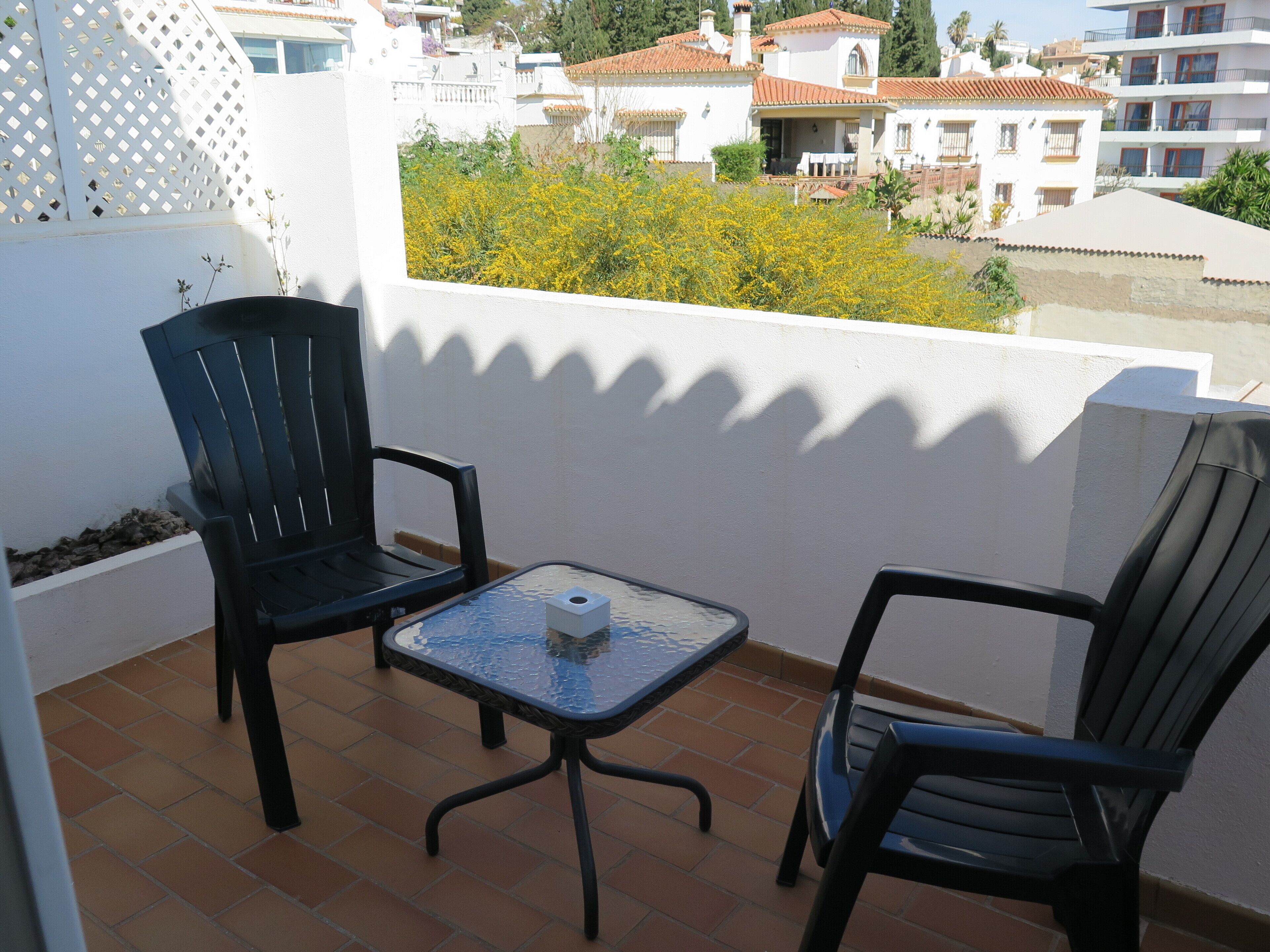 Un balcón con dos sillas negras y una pequeña mesa, con vistas a una zona residencial de edificios blancos y techos de tejas rojas.