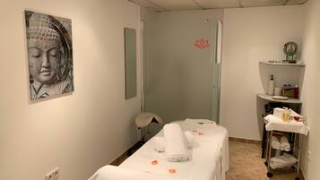 A spa room with a massage table, a wall-mounted mirror, a shelf with toiletries, and a Buddha painting.