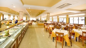 A buffet-style restaurant with multiple dining tables and chairs, a central serving area with food displays, and large windows providing natural light. - Hotel Monarque Fuengirola Park (Fuengirola)