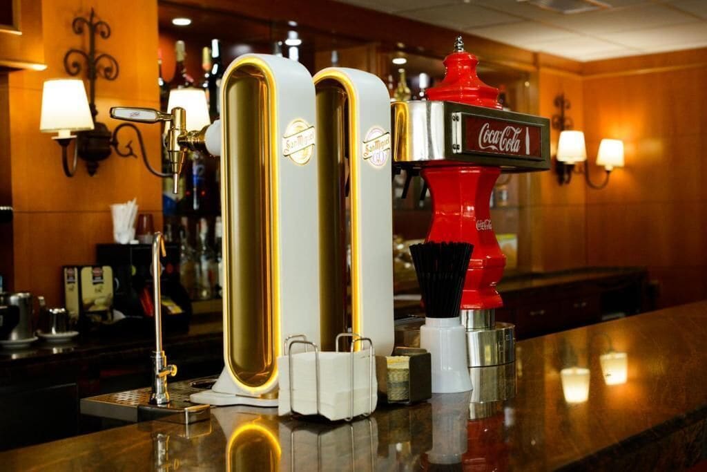 A bar area with a Coca-Cola branded machine, two tall, cylindrical dispensers, and a coffee machine.