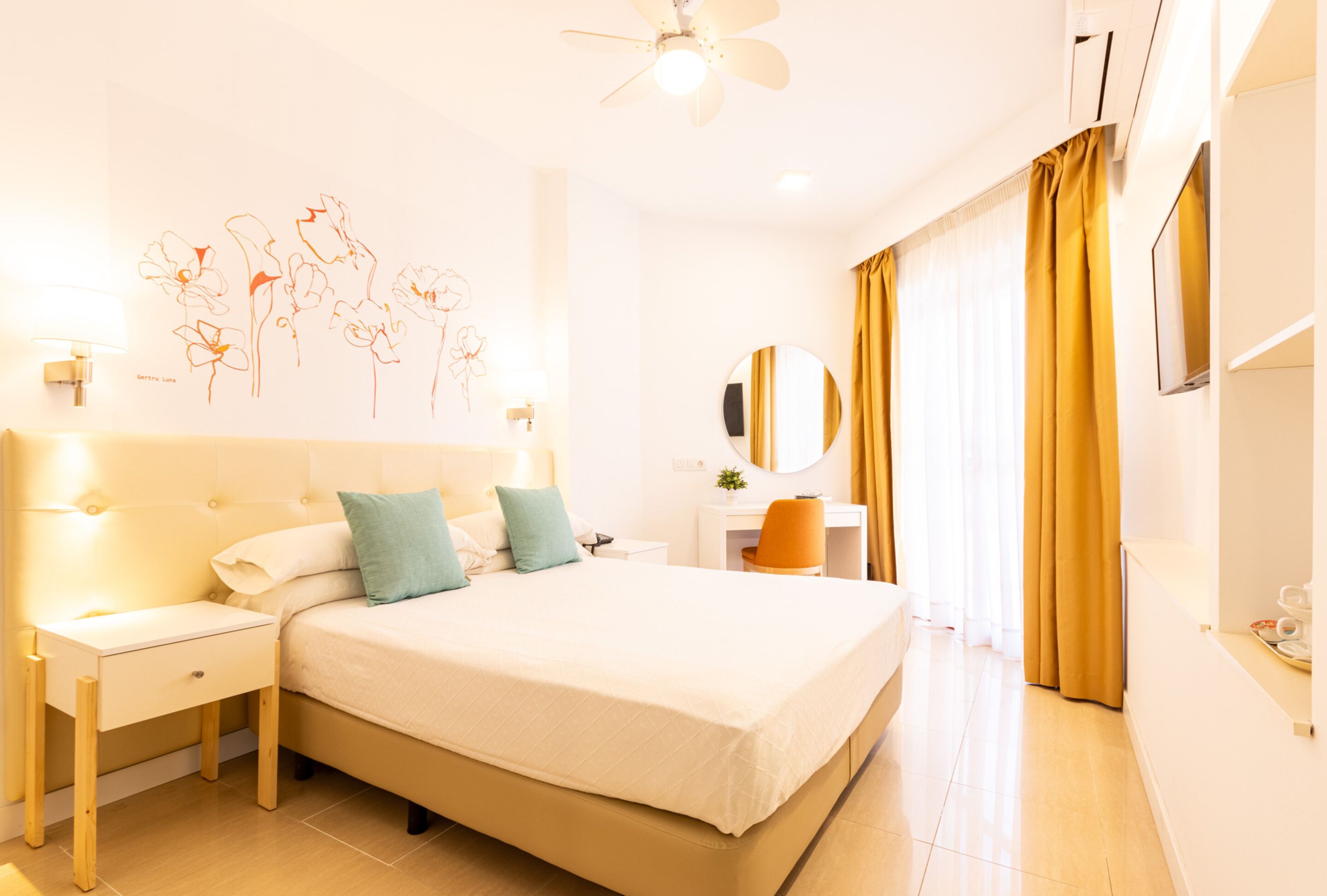 A hotel room with a large bed, bedside tables, a desk, a chair, a ceiling fan, a mirror, and curtains.