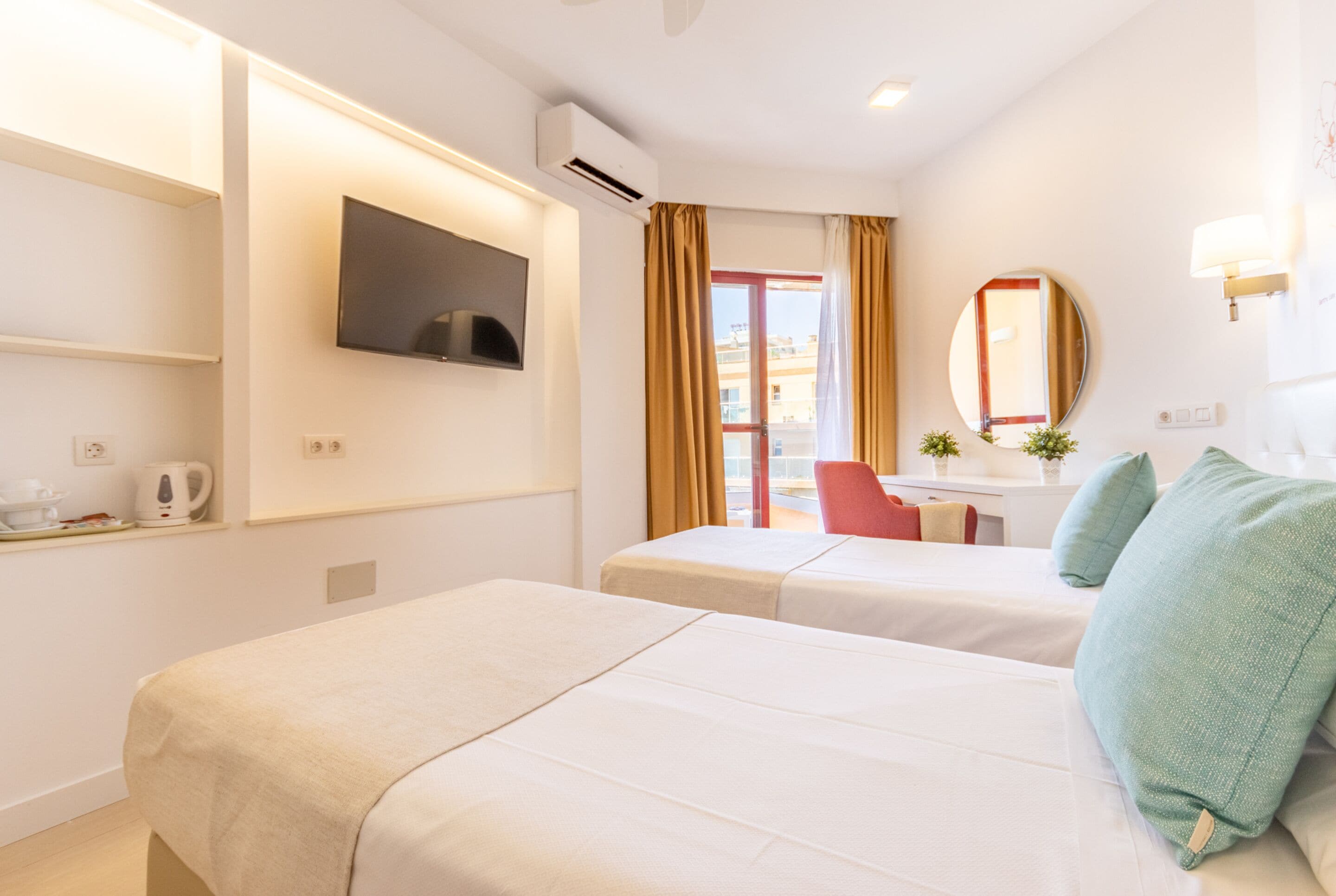 A hotel room with two beds, a TV mounted on the wall, a small desk with a chair, and a window with curtains.