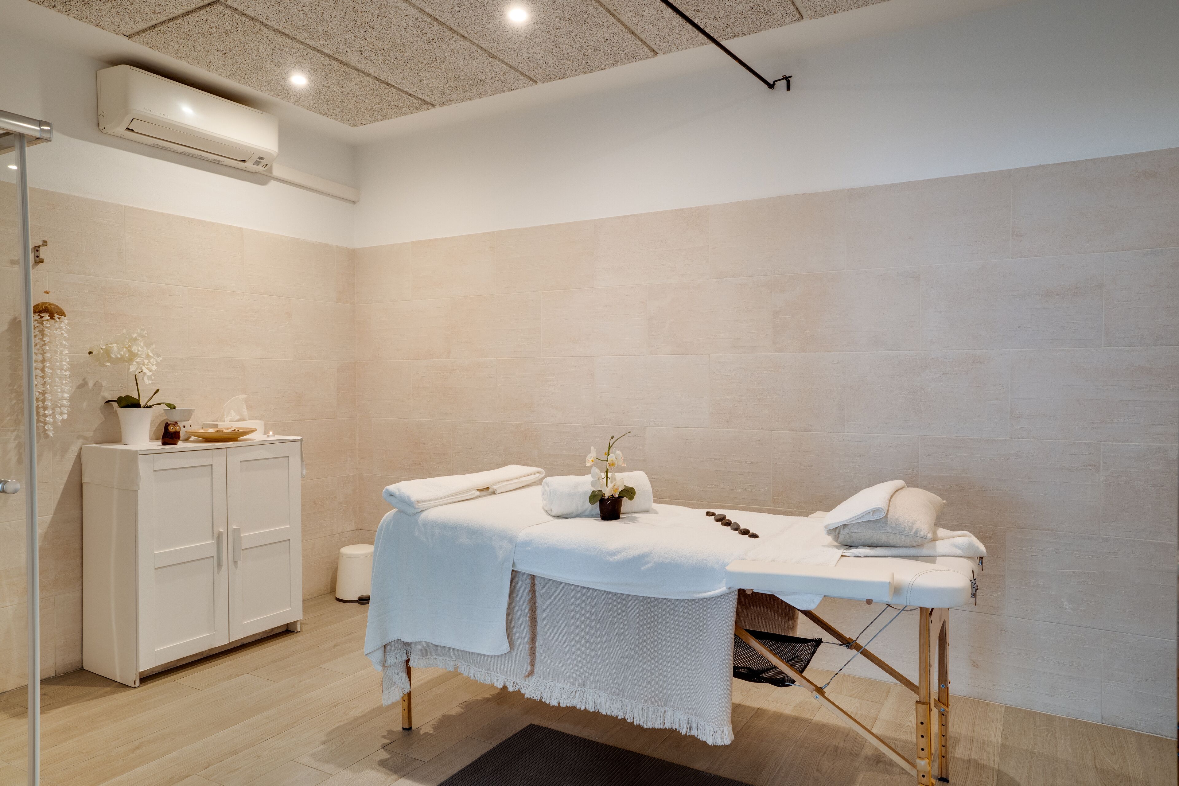 Een schone, moderne spa-ruimte met een massagetafel, witte handdoeken en een kastje met decoratieve objecten.
